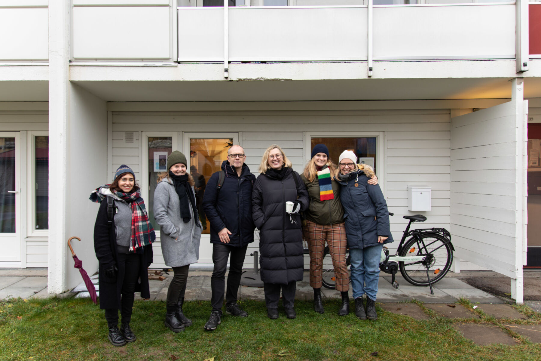 Seks personer står sammen foran et inngangspart på et hvitt trehus. De ser mot kamera og smiler. Foto.