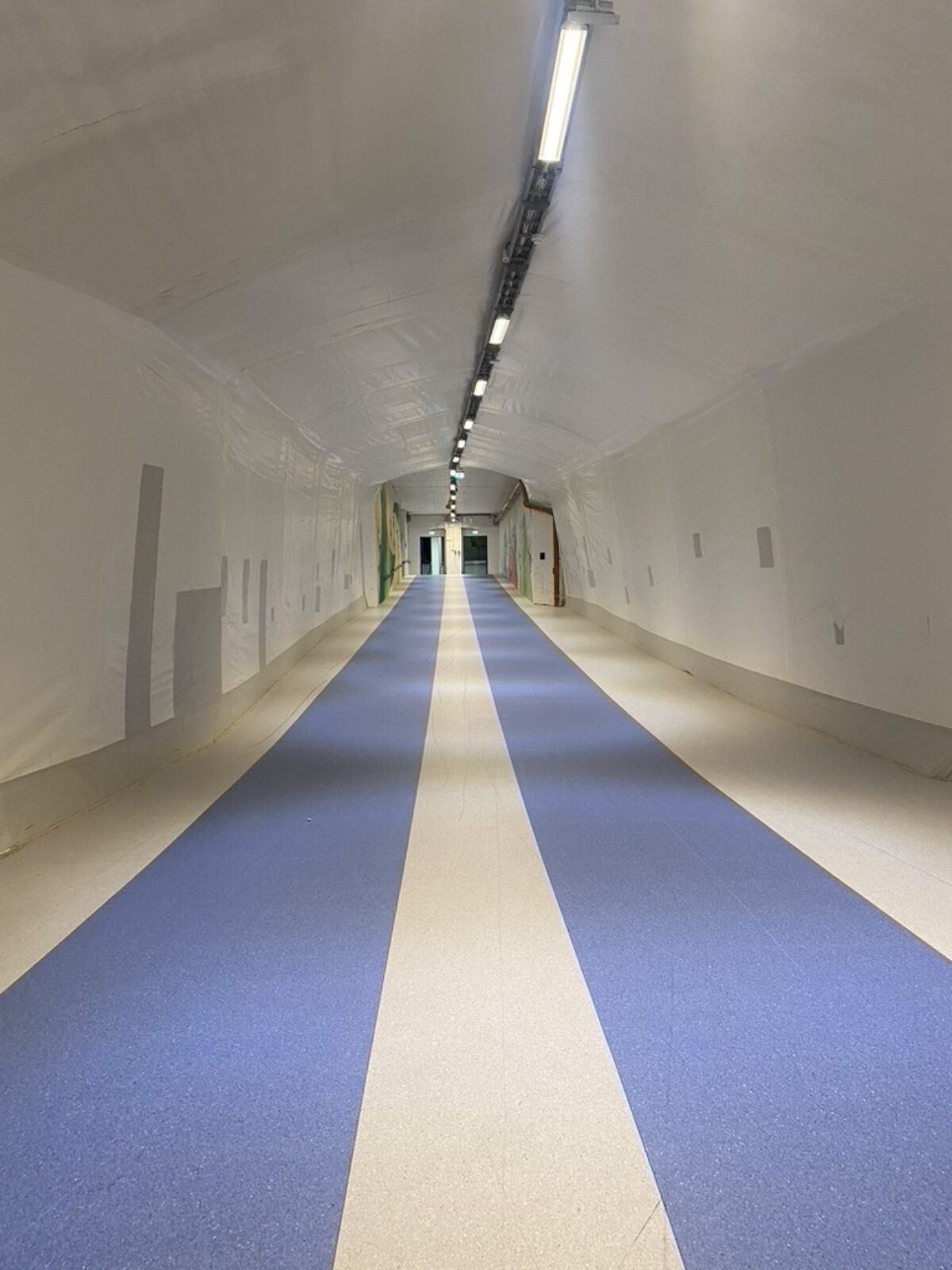 En lang korridor i idrettshall, to blå løpestriper på gulvet. Foto.