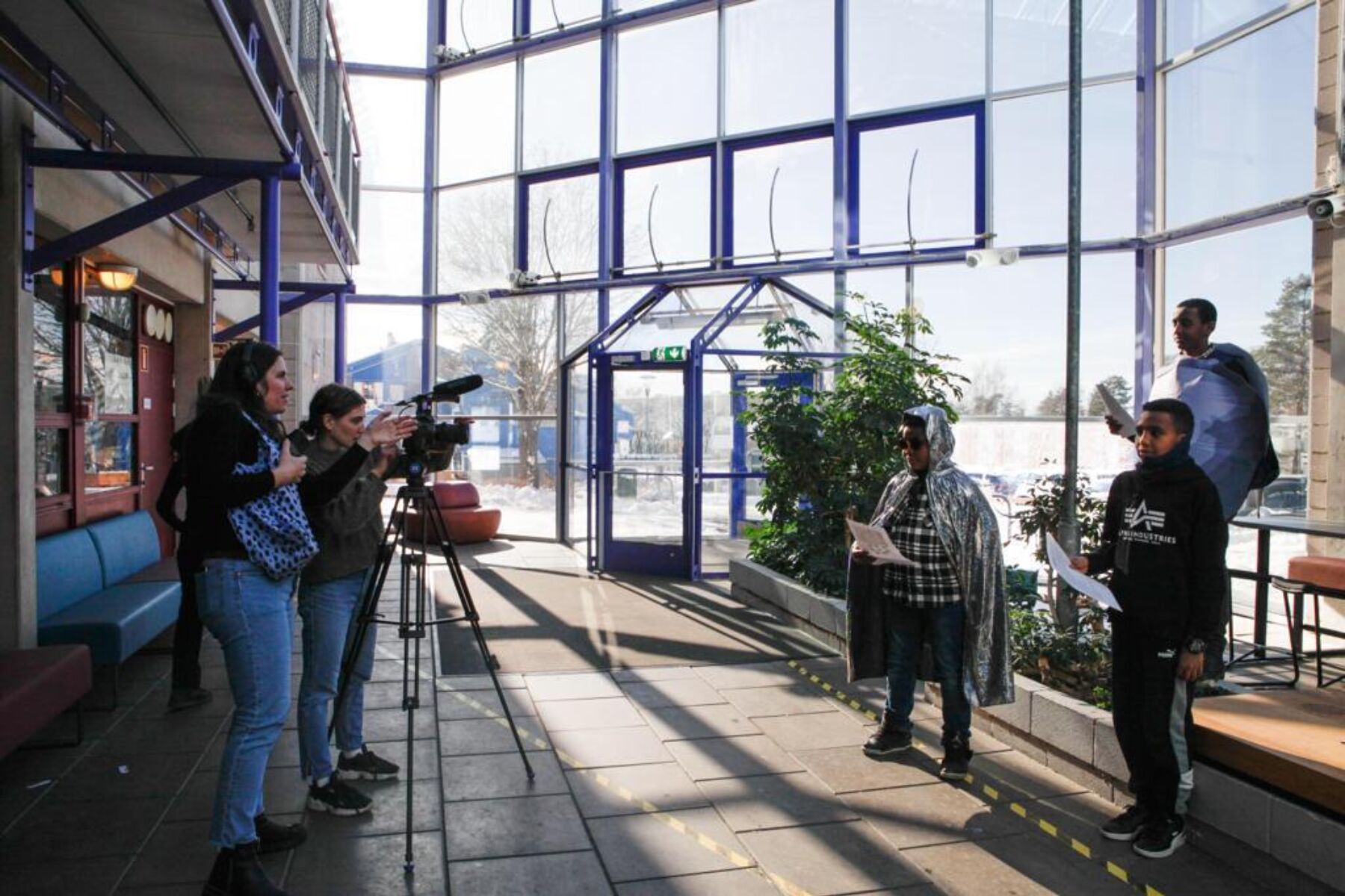 To personer til venstre i bildet står med filkamera som peker mot tre barn på høyre side. De står inne i et glassbygg. Foto.