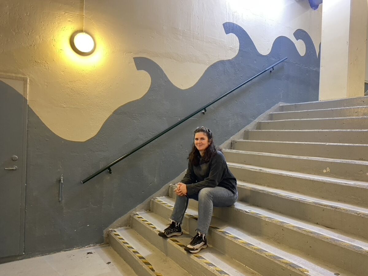 Person sitter i trapp med bølge malt på vegg i bakgrunn. Foto.
