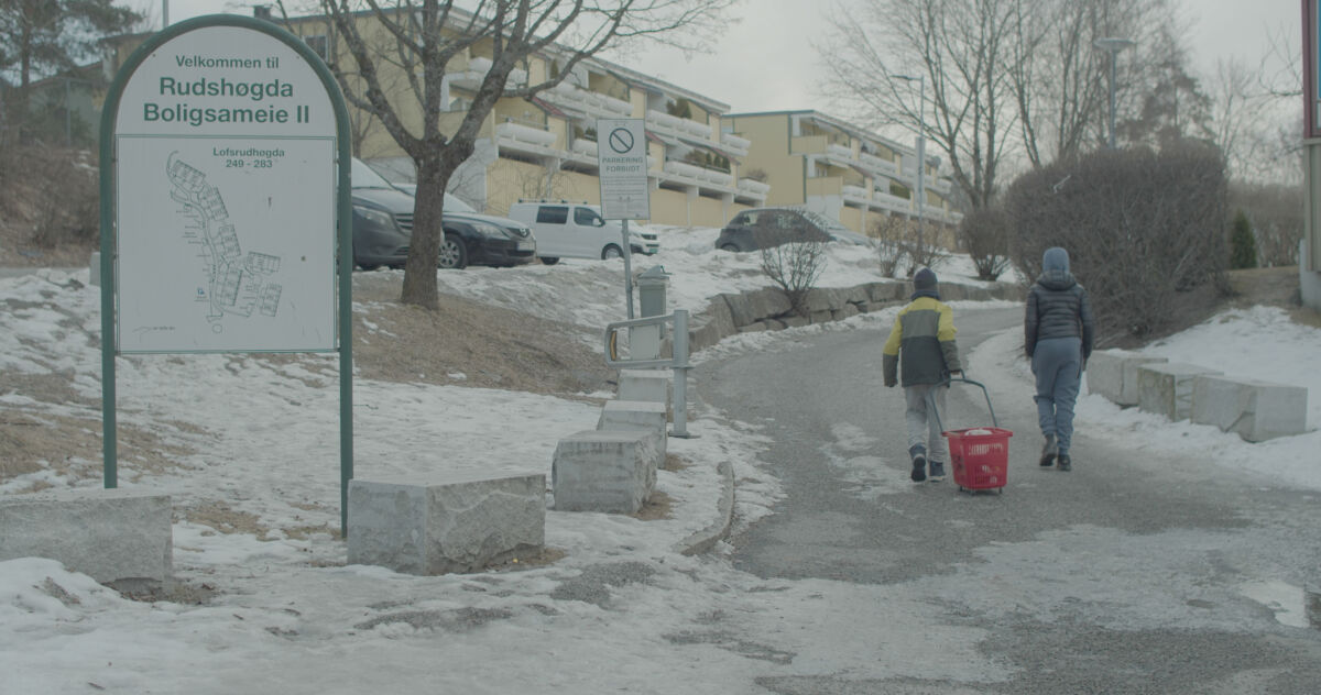To personer går opp en vei mellom boligblokker i vinterlandskap. Foto.