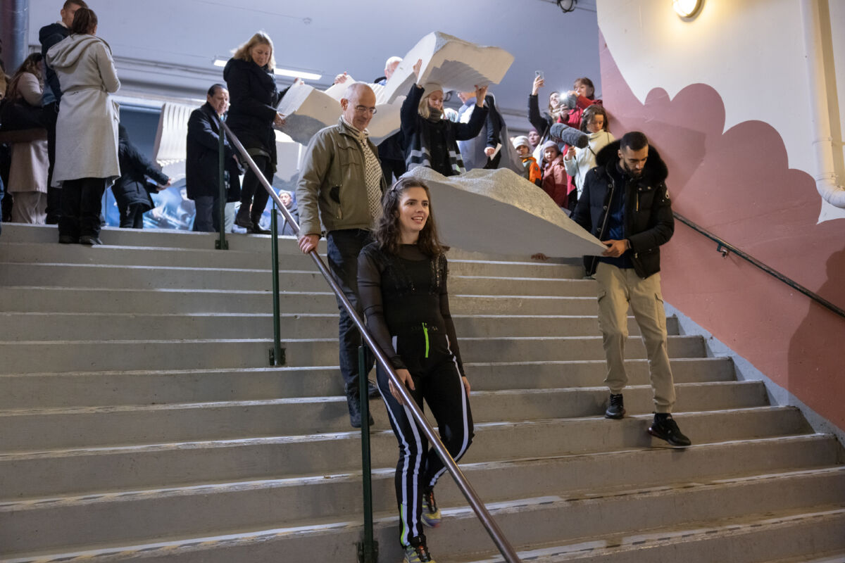 Flere personer går sammen ned en trapp mens de bærer en stein i flere deler, som en del av en forestilling. Foto.
