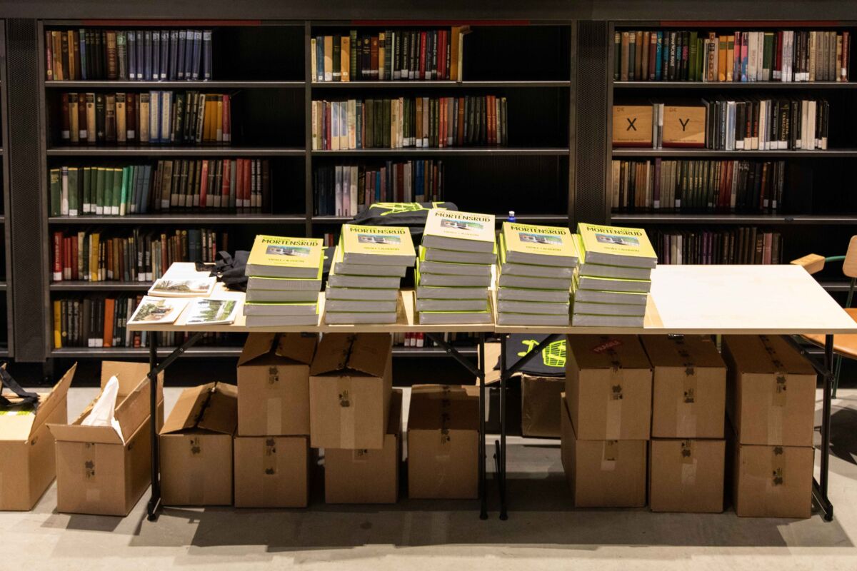 Bøker og kasser med bøker stablet på og under et stort bord i et bibliotek. Foto
