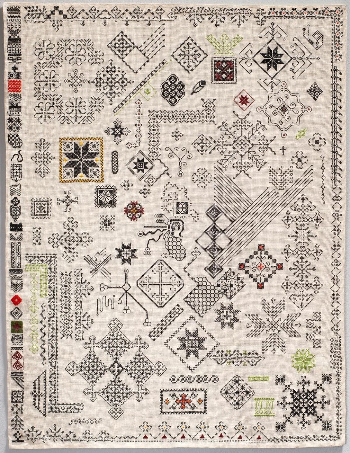 Brodert tekstil i hvitt med ulike mønstre og motiver i svart, rødt og grønt. Foto av tekstil.