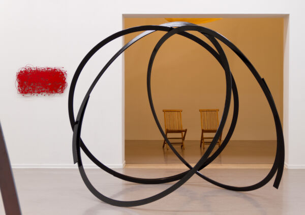 En svart, sirkulær skulptur som står på gulvet. I bakgrunnen er to trestoler og et rødt kunstverk på veggen. Foto.