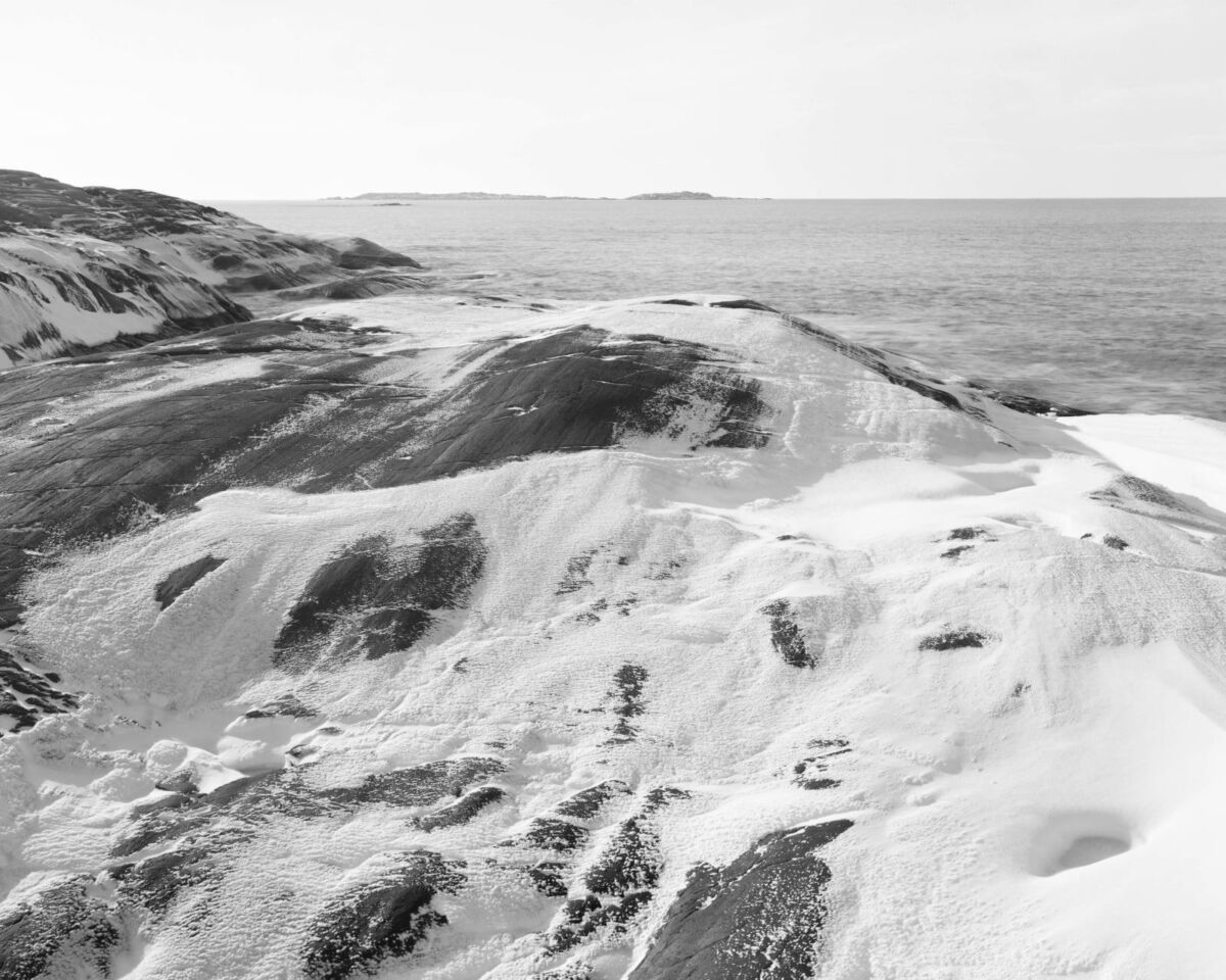 Svart-hvitt foto av berg dekket med snø i vannkanten. I bakgrunnen ligger havet. Foto.