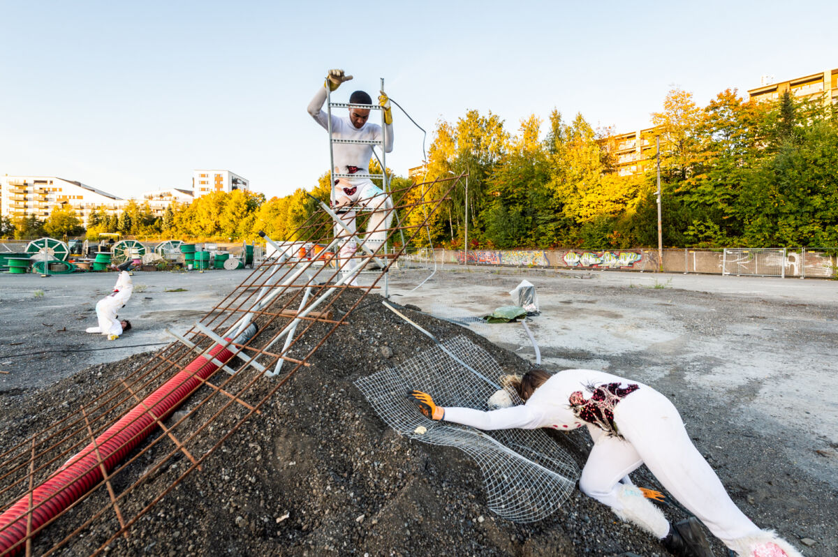 To mennesker klatrer opp mot en liten steinhaug på en parkeringsplass. I bakgrunnen ligger en person med føttene opp mot himmelen. Foto.