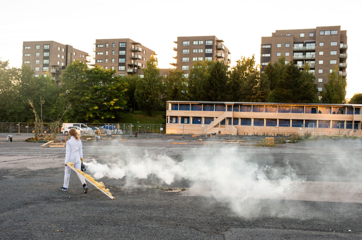 En person trekker en planke etter seg på en. Det kommer røyk fra et apparat som personen bærer på i høyre hånd. I bakgrunnen står fire høyblokker med trær foran. Foto.