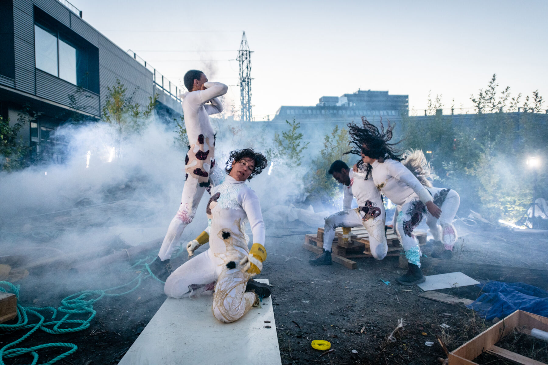 Fire mennesker danser eller beveger seg på bakken utendørs. Rundt dem er søppel, røyk og materiale. Foto.