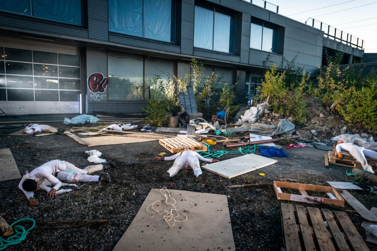 Fire mennesker ligger på bakken utendørs. Rundt dem er søppel og materiale. Foto.