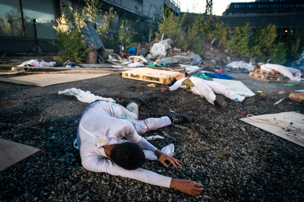 Fire mennesker ligger på bakken utendørs. Rundt dem er søppel og materiale. Foto.