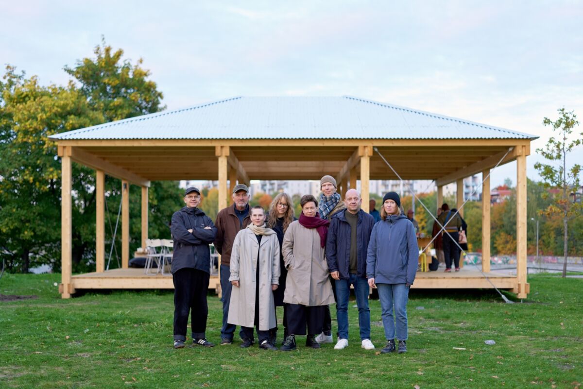Åtte mennesker står samlet foran en paviljong i tre. Foto.