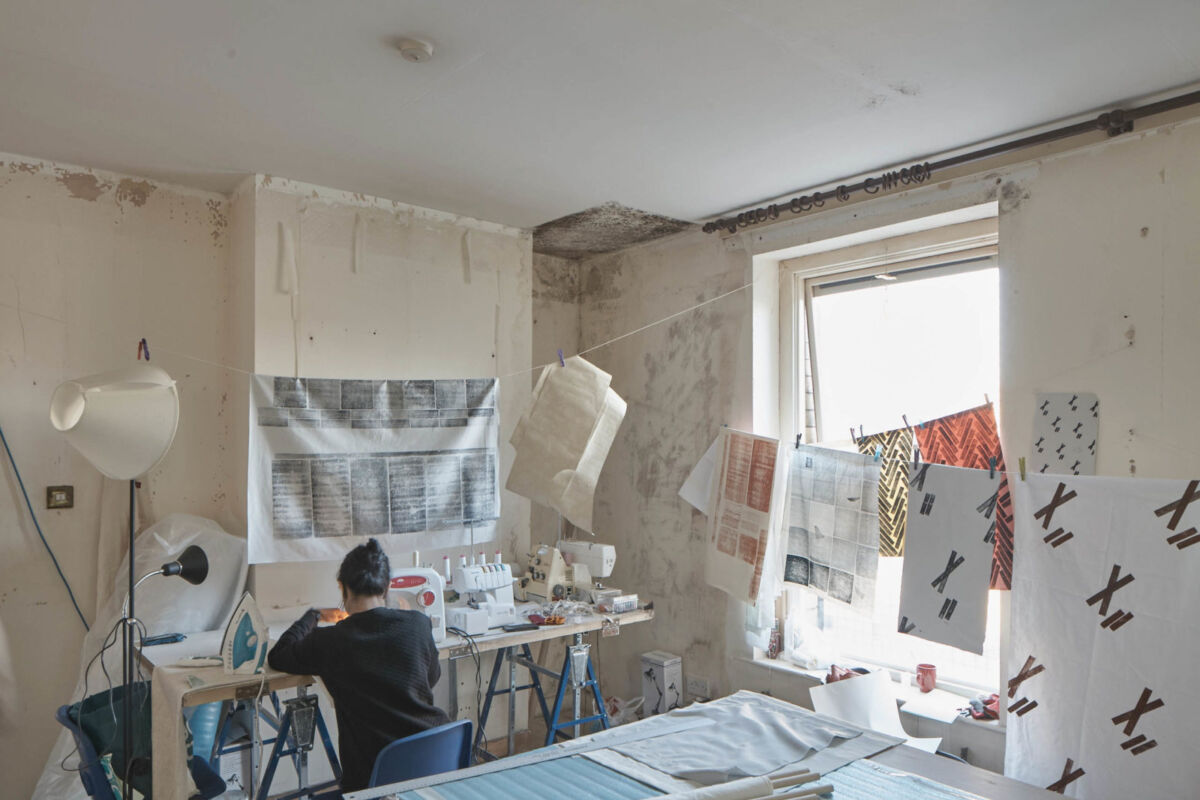 En person sitter ved en symaskin og jobber. I resten av studioet henger tekstil med ulike mønstre. Foto.