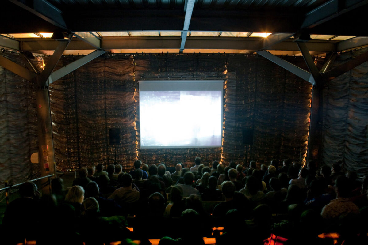En mørk kinosal hvor skjermen lyser og mange mennesker sitter i publiku. Foto.