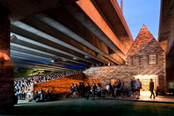 Mange mennesker sitter i et atrium på rekke og rad under en bro. Foto.