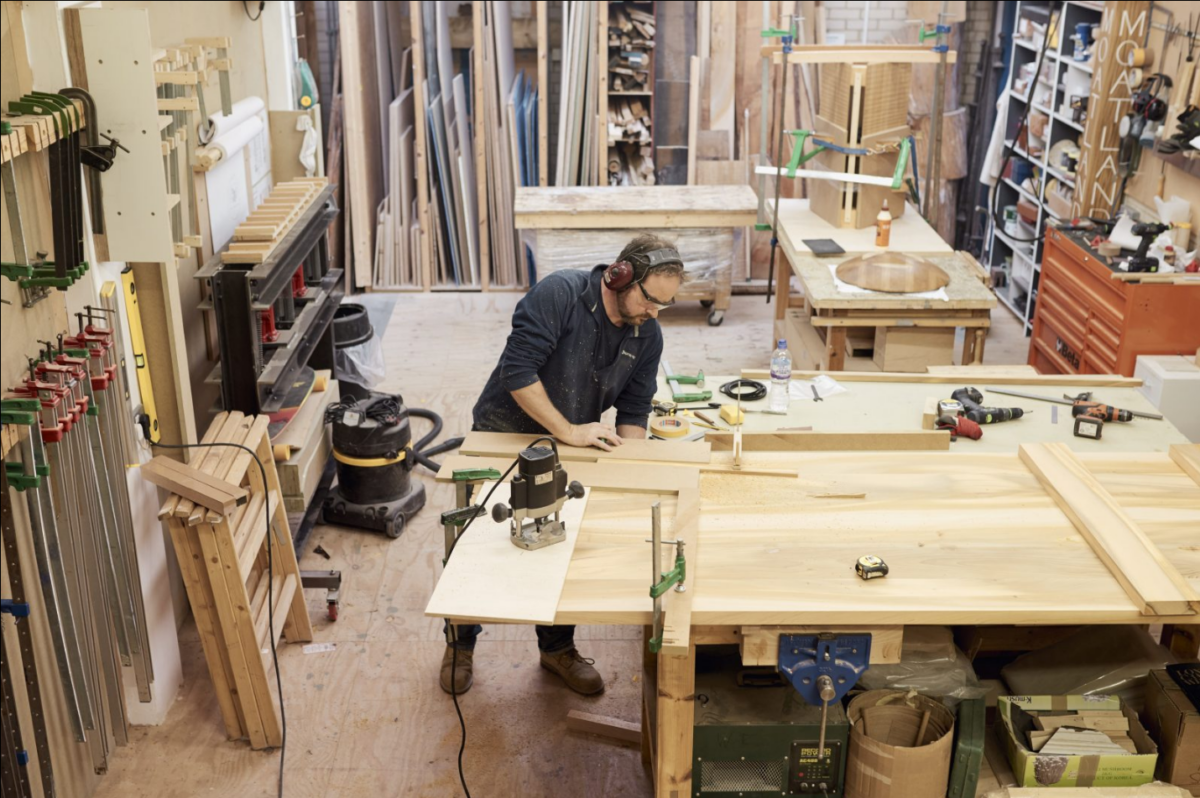 En person jobber med treverk på et lyst verksted. Det henger, står og ligger verktøy og materiale rundt. Foto.