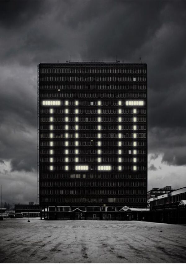 Svart-hvitt foto av høyblokk med mange vinduer. Noen av vinduene lyses opp, slik at ordet "TILLIT" står skrevet på bygningen. Foto.