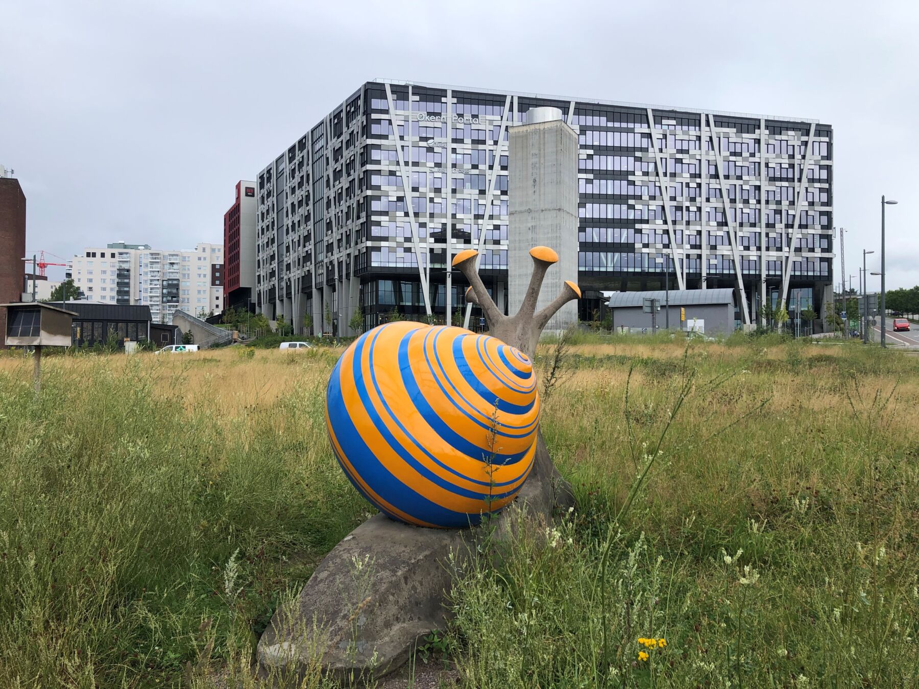 Skulptur av snegle i grønn eng foran næringsbygg i bakgrunnen. Foto.