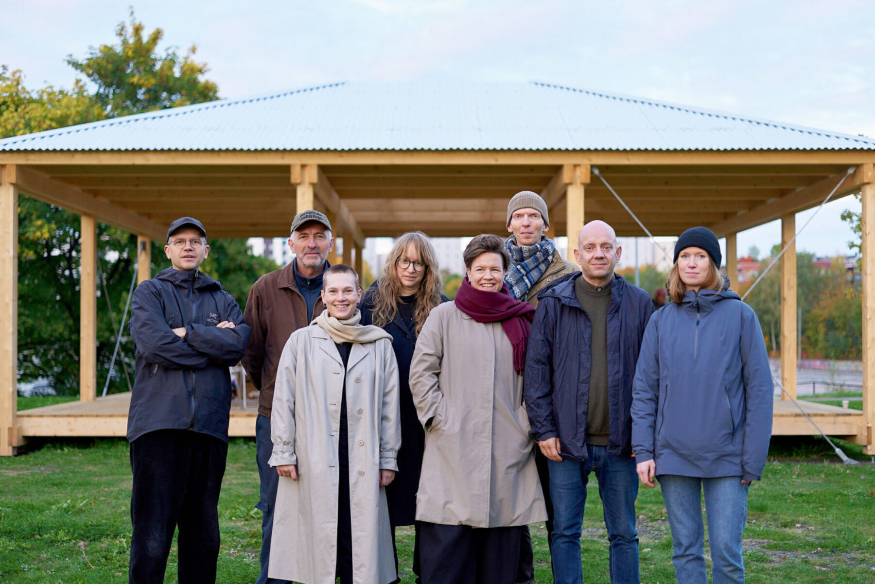 Åtte personer står utendørs. De ser mot kamera. I bakgrunnen er en paviljong i tre. Foto.