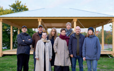Åtte personer står utendørs. De ser mot kamera. I bakgrunnen er en paviljong i tre. Foto.