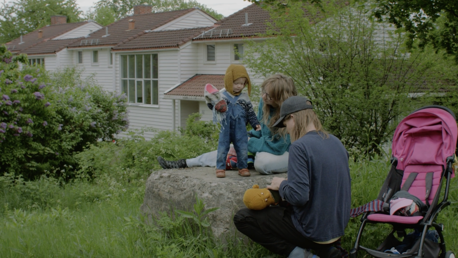 To voksne er utendørs i en hage med et barn som står mellom dem på en stein. Foto.