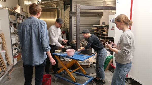 Fire personer stående rundt et arbeidsbord i et verksted. De lager murstein. Foto.