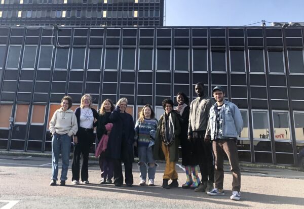 Ni mennesker fotografert forfra. De ser alle mot kameraet. De står på en parkeringsplass, og bak dem er en stor bygning. Foto.