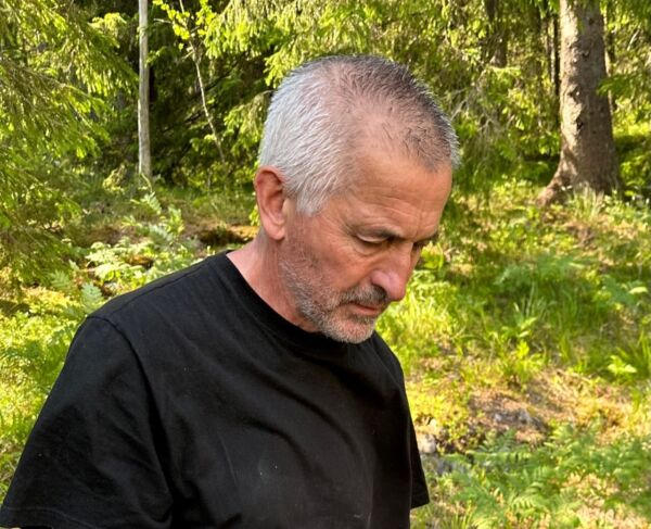 Person står foran kamera og ser ned mot bakken. I bakgrunnen er det grønn skog. Foto.