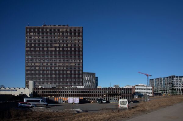 Høyblokk med mange vinduer. I bakgrunnen er himmelen blå, og i forgrunnen er en parkeringsplass med biler. Foto.