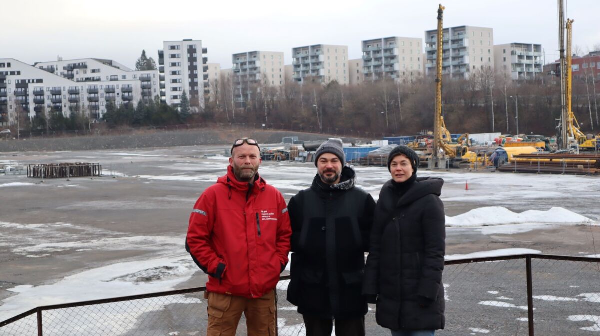 Tre personer står samlet utendørs. I bakgrunnen er en stor plass og mange boligblokker. Foto.