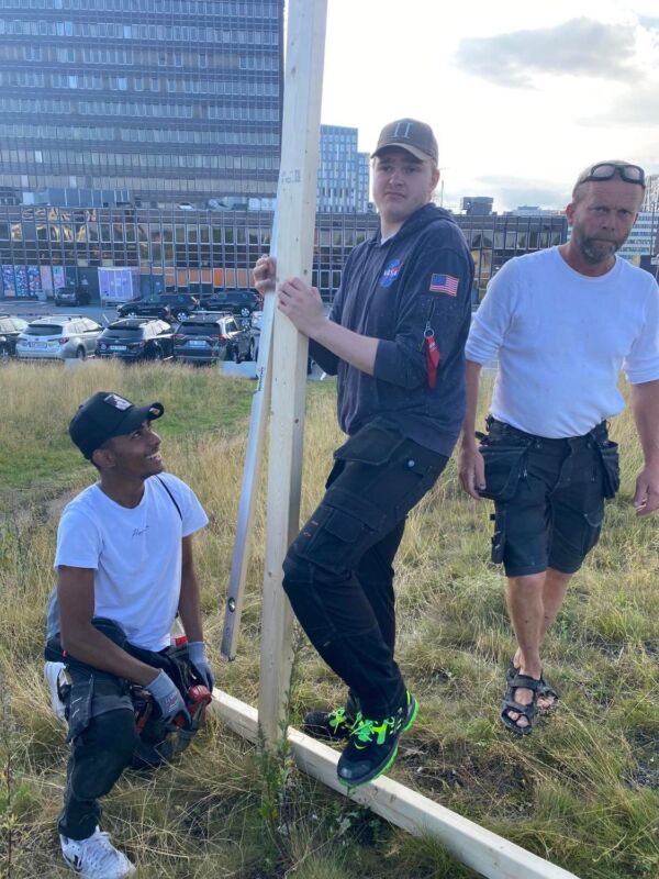 Tre personer jobber med treplanker utendørs i gresset. Foto.
