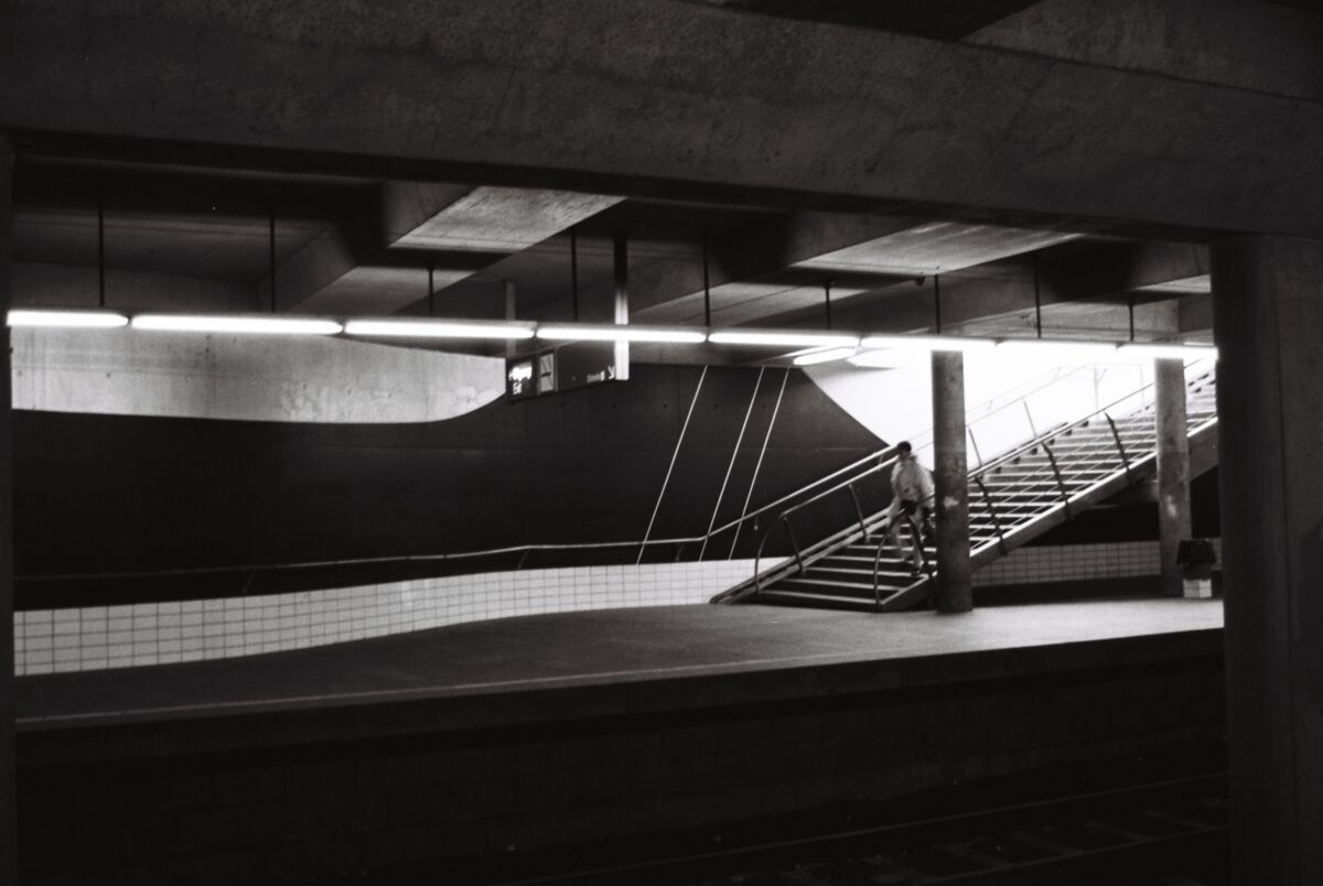 Person går nedover en trapp og ned på en T-baneperrong. Det er mørkt i rommet og på veggen er et veggmaleri. Analogt foto i sort-hvitt.