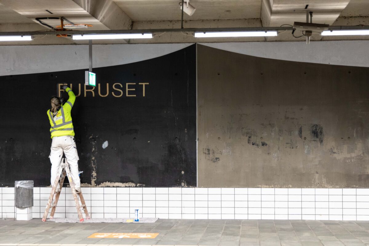 Person maler på et veggmaleri med teksten Furuset, innendørs på en t-baneplattform. Foto.