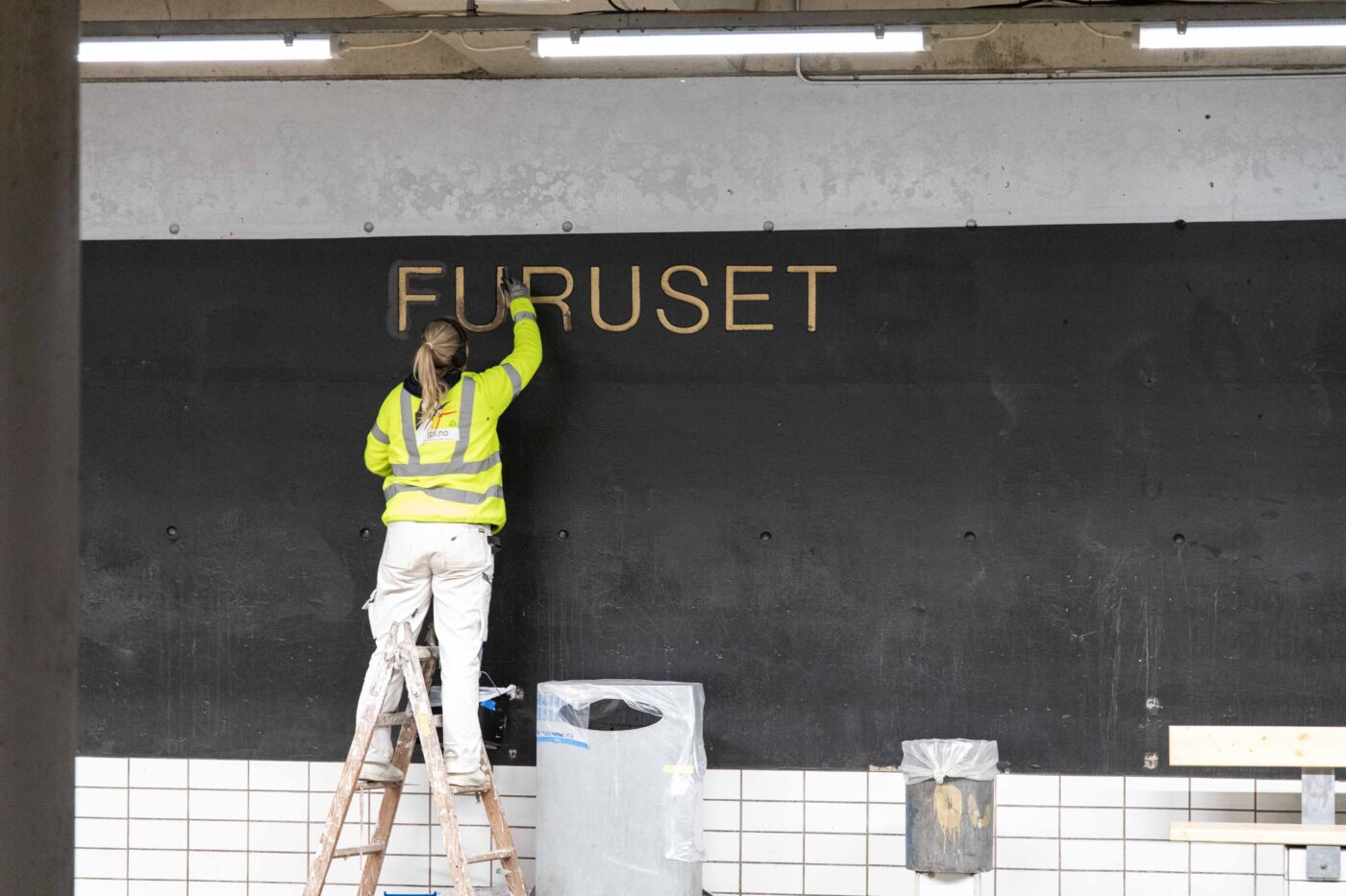 Person maler på et veggmaleri med teksten Furuset, innendørs på en t-baneplattform. Foto.