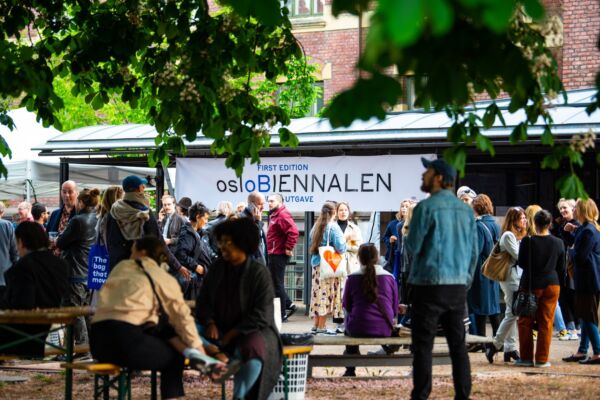 Mange mennesker samlet på et grønt område. Det henger et banner med teksten "osloBIENNALEN" fra et tak. Foto.