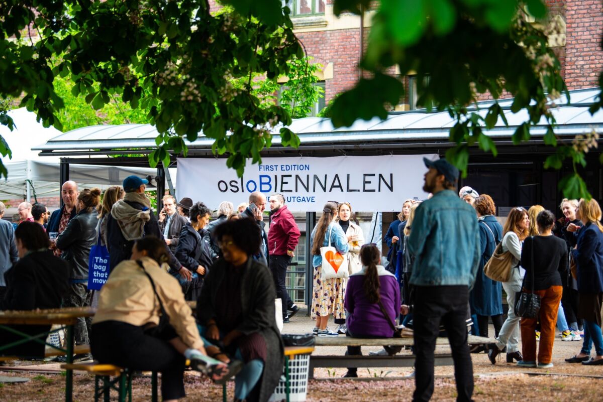 Mange mennesker samlet på et grønt område. Det henger et banner med teksten "osloBIENNALEN" fra et tak. Foto.