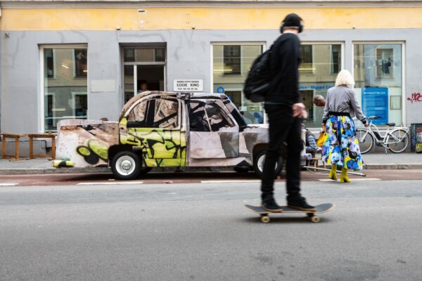 En bil dekorert med graffiti står parkert i sykkelfeltet utenfor et gult og grått bygg. En person går i nærheten, mens en annen person skater forbi i forgrunnen. Foto.