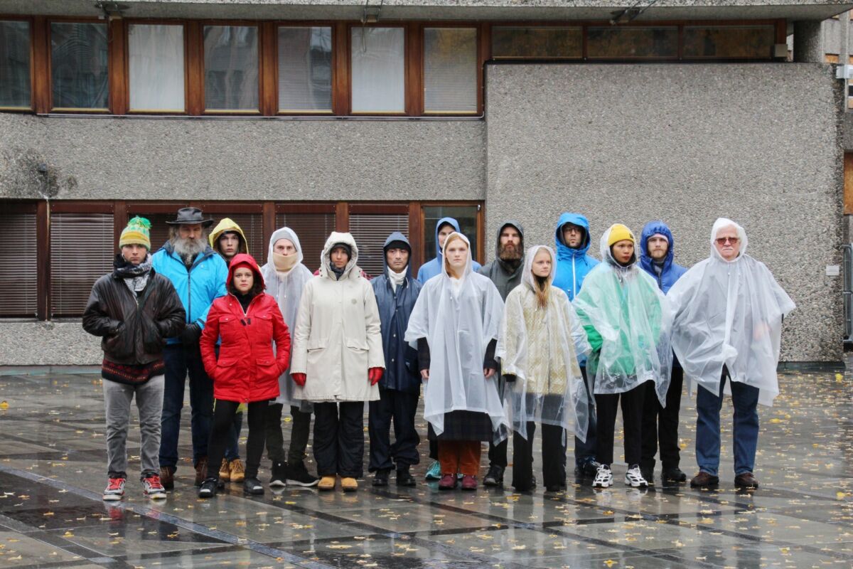 En gruppe mennesker står rett opp og ned i en gruppe foran et brutalistisk bygg. De ser rett frem og har på seg regntøy. Foto.
