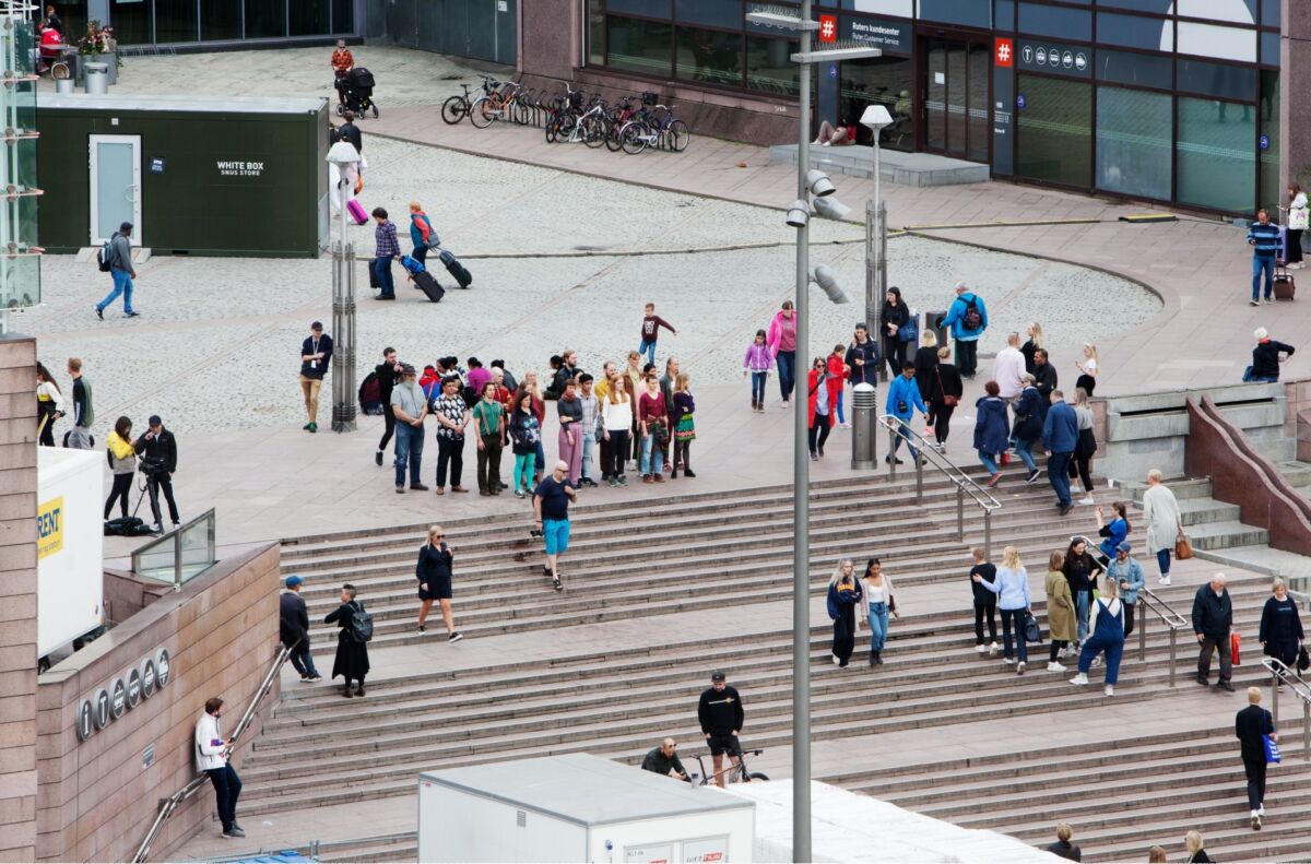 Bilde tatt ovenfra av en gruppe mennesker som står og ser rett frem. Rundt dem beveger mennesker seg. Foto.