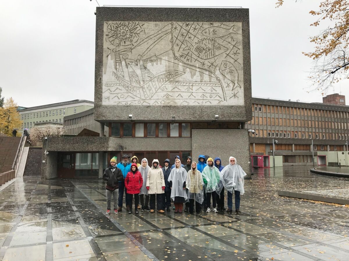 En gruppe mennesker står rett opp og ned i en gruppe foran et brutalistisk bygg. De ser rett frem og har på seg regntøy. Foto.