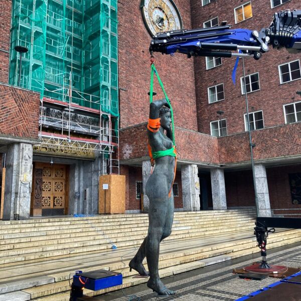 En stor skulptur av en kvinne sett løftes med lift foran et stort murbygg. Foto.