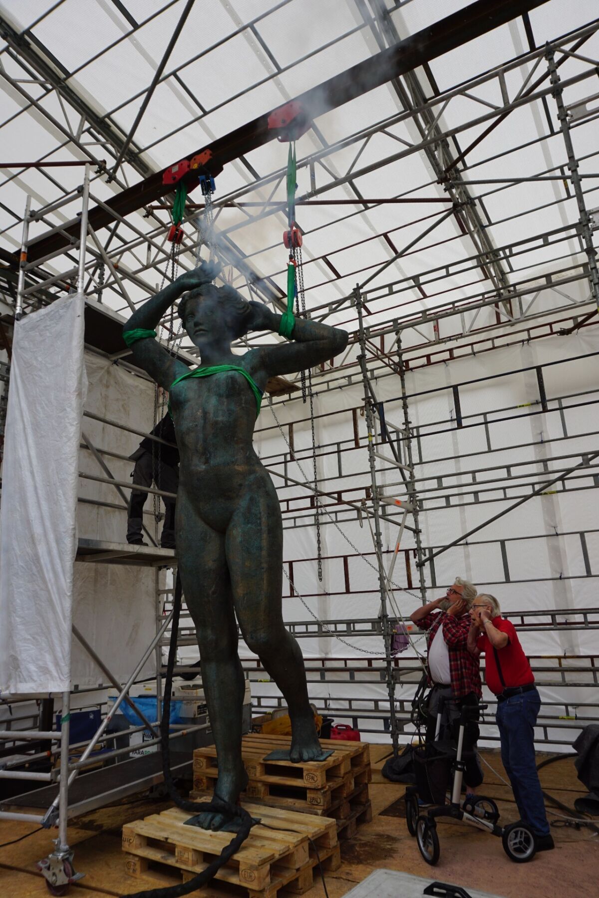 En stor skulptur av en kvinne sett forfra. Skulpturen står inne i et telt med stillaser rundt og to personer ser opp på den og måper. Foto.