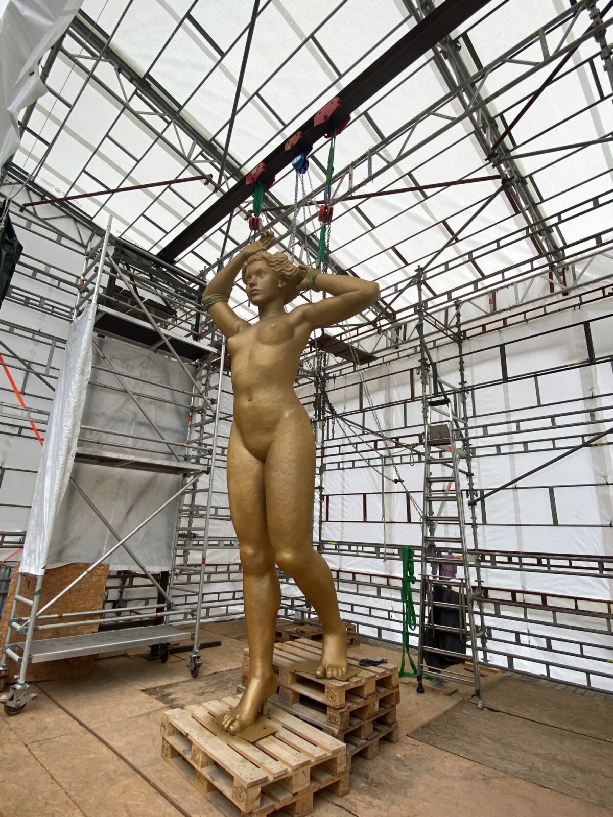 En stor, gullforgylt skulptur av en kvinne. Skulpturen står inne i et telt med stillaser rundt. Foto.
