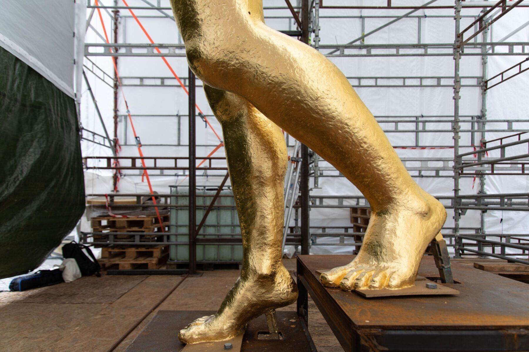 Nærbilde av to føtter på en gullforgylt skulptur. Skulpturen står inne i et telt med stillaser rundt. Foto.