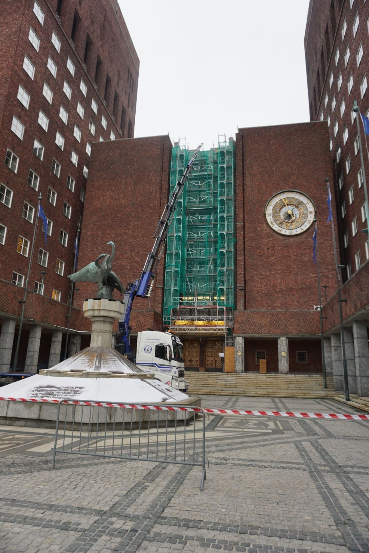 En stor murbygning omkranser et torg. I midten løftes en integrert skulptur ned fra veggen med lift. Foto.