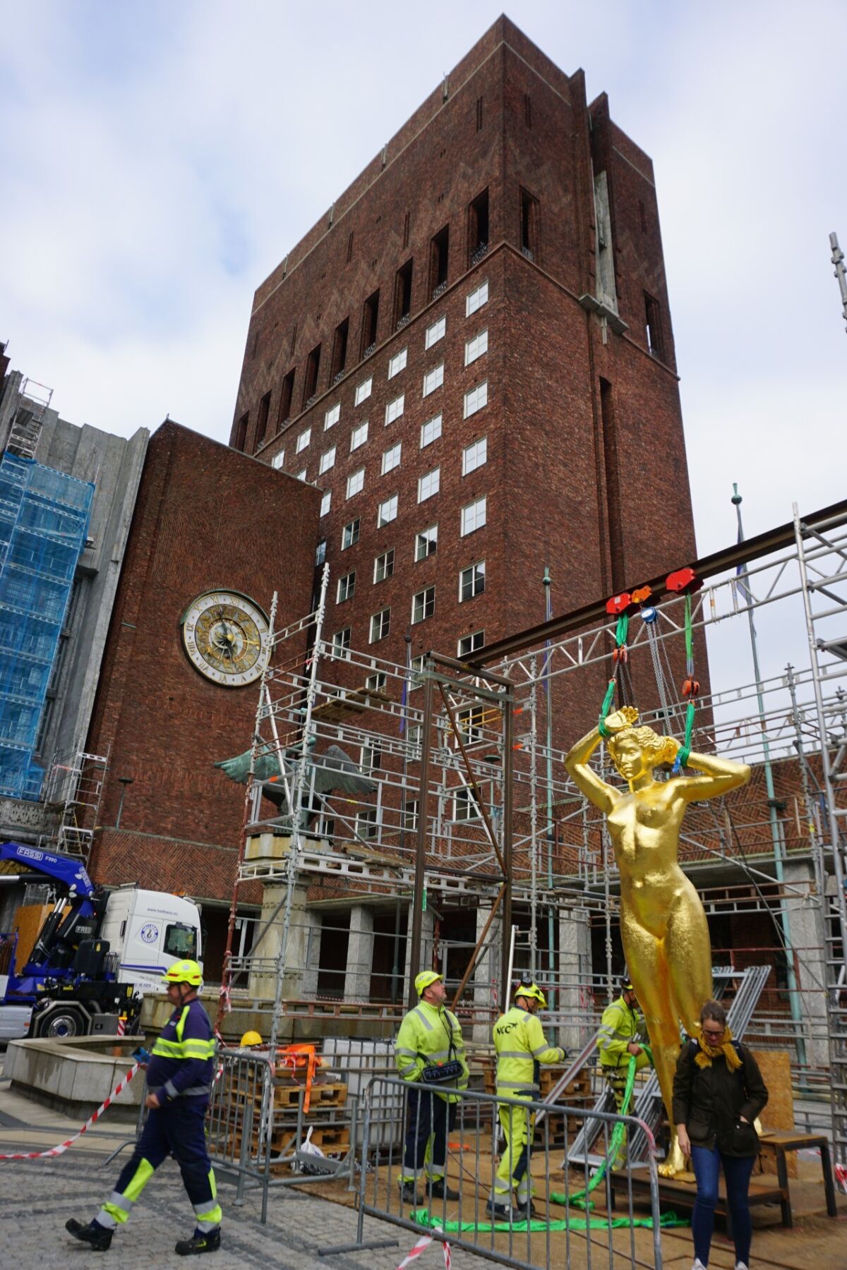 En stor, gullforgylt skulptur av en kvinne er i gang med å heises opp. I bakgrunnen er en stor murbygning. Foto.