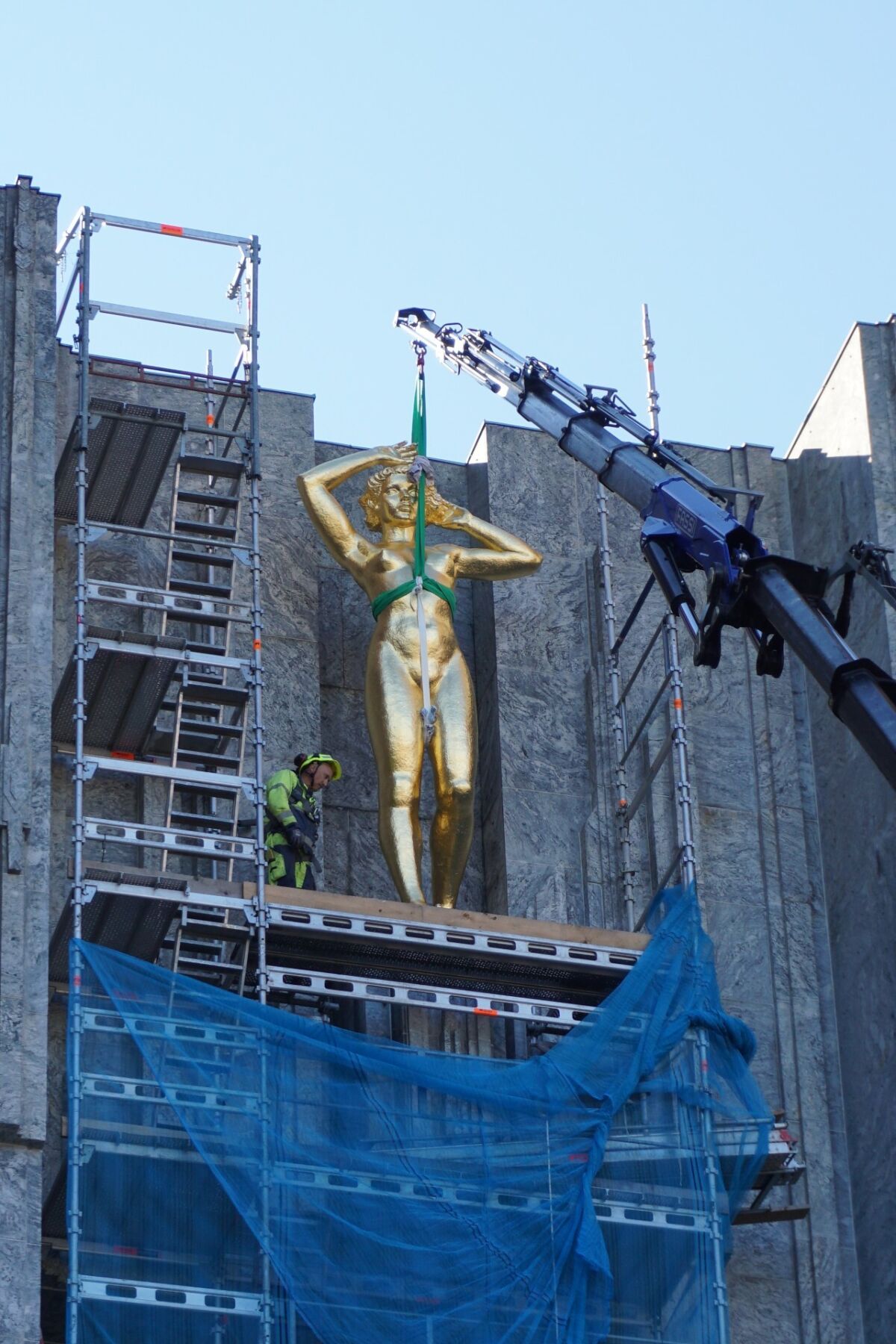 En større gullforgylt statue av en kvinne løftes ved hjelp av lift opp foran en murbygning. Foto.