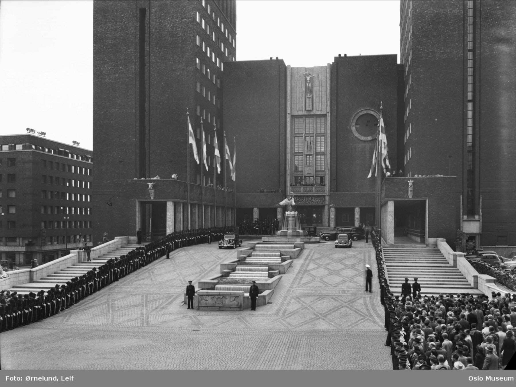 Svart-hvit foto av en stor murbygning med en åpen plass foran og mange mennesker som ser på. Bildet er tatt fra åpningen av Oslo rådhus i 1950. Foto.