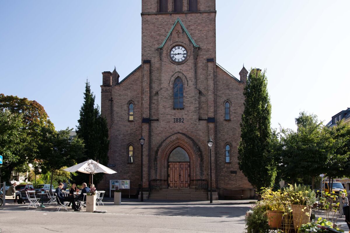 Kirke i rød teglstein i bakgrunn, byroms plass med sittegrupper og beplantning. Foto