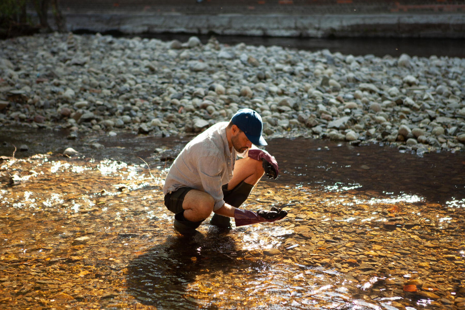 En person sitter på huk i en elv og plukker opp noe fra vannet. Foto.